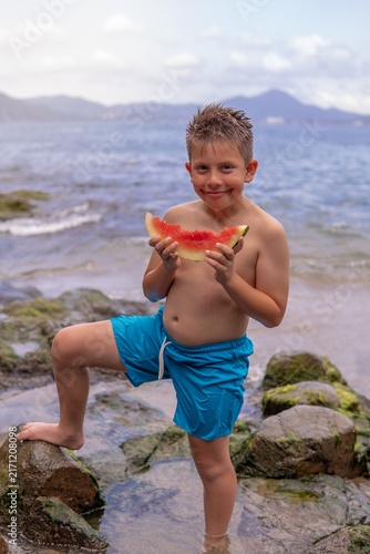 niño pequeño rubio parado sobre arena y rocas en el mar en una playa comiendo un rebanada de sandia dulce y deliciosa al aire libre en la naturaleza disfrutando de un hermoso dia 