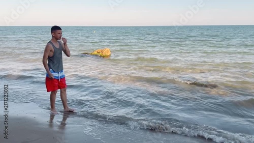 Energetic teenage boy laughs while catching and tossing a water ball between his legs on the beach during a sunny summer holiday. High quality FullHD footage
