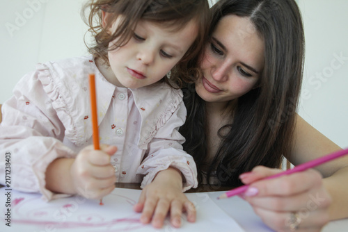 Bild auf Leinwand A little girl focuses on drawing with colored pencils while her mother sits by her side supporting her creative activity at a table at home
