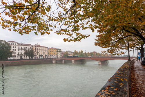 Fotografie Wide view of autumn tree foliage framing river embankment and historic bridge in
