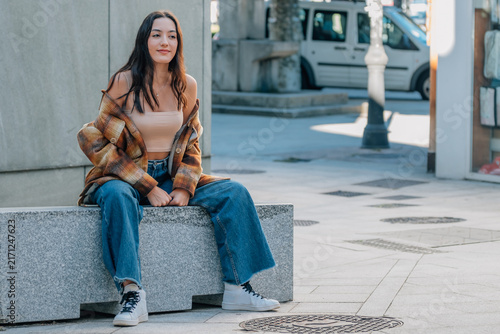 Young woman with long brown hair sitting outdoors on a stone bench, smiling and looking away, wearing a plaid flannel shirt jacket, autumn urban lifestyle portrait