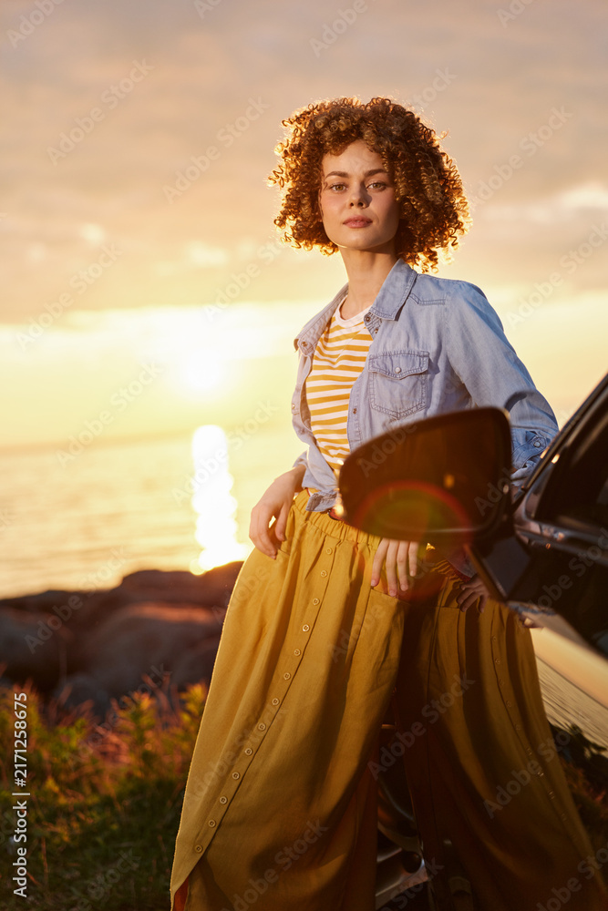 Naklejka premium A woman with curly hair stands beside a car at sunset, wearing a striped shirt and denim jacket, capturing a relaxed lifestyle moment. Concept: casual confidence and outdoor freedom.