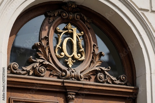 Ornate wooden door detail with carved embellishments and golden letter d. Arched window above features dark reflection, architectural elements show classic European style. Detailed facade.