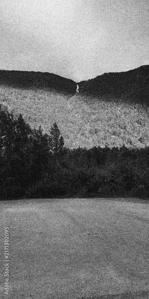 Fototapeta premium Distant Narrow Waterfall Between Forested Hills in Heavy Grain Black and White Landscape
