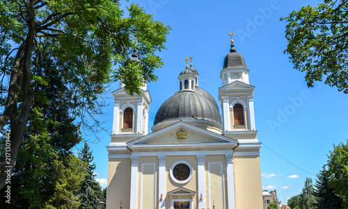 Neoclassical peach facade of the Holy Spirit Cathedral in Chernivtsi, Ukraine, framed by summer trees under a blue sky, featuring twin bell towers and a central dark grey dome