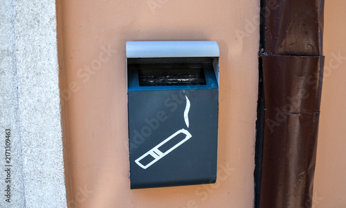 Dark grey, wall mounted public ashtray on a peach colored exterior wall with a brown downspout, representing urban waste management and public hygiene