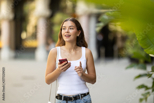 Young smiling girl with phone walks along city street, walks from work to home. Pedestrian in summer European capital, person in city, city bustle.