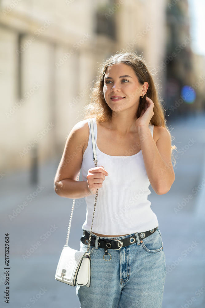 Naklejka premium Young woman smiling and walks along sunlit street, walks around city, spends time in public urban space. Person and megalopolis..