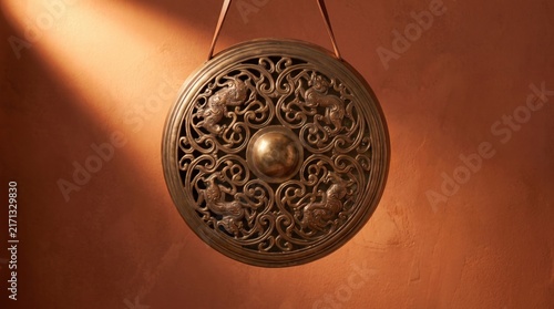 An ornate bronze gong hangs against a warm textured wall illuminated by soft golden light beams.