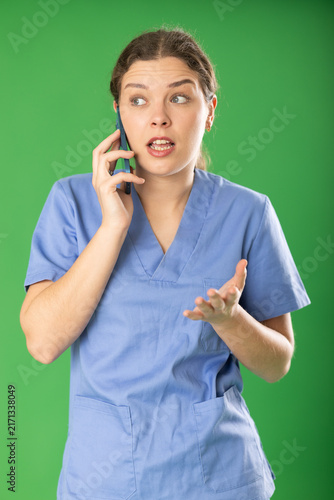 Young professional female doctor wearing blue medical clothing convincingly and emotionally talking to patient by smartphone standing against green screen