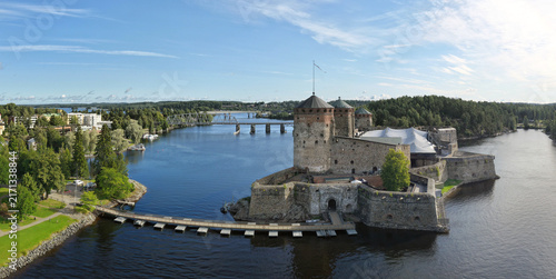 Savonlinna, Finland - 14 August 2026: Aerial view of Olavinlinna Castle surrounded by water, lush green forests, and a bridge in the background under a clear blue sky.