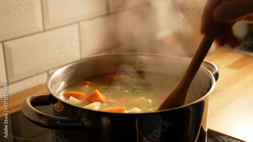 Close-Up Of A Pot Of Simple Soup Being Stirred On A Home Stove Featuring Fresh Vegetables And Steam Rising For A Warm, Comforting Meal