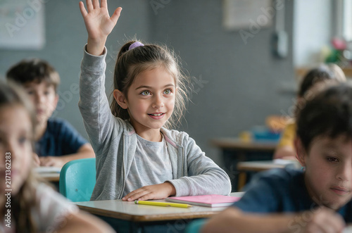 Young girl raises hand in classroom, smiling brightly. Other children sit quietly, focused on lesson. Bright lighting shows cheerful, engaged learning environment. Ideal for education, school