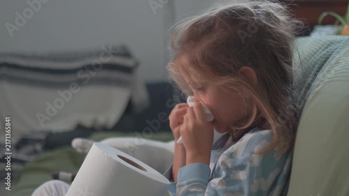 A young girl sits in her living room, feeling sick and holding a tissue. She looks around, seeking warmth and care while wrapped in a blanket on a soft couch.