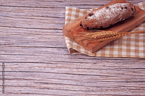 Fresh sourdough rests on wooden board, dusted with flour. Warm bread sits atop rustic cutting board. Wheat stalks lie beside it, woven mat below. Subtle shadow hints at soft, cozy kitchen light