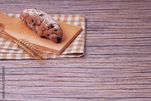 Fresh sourdough rests on wooden board, dusted with flour. Warm bread sits atop rustic cutting board. Wheat stalks lie beside it, woven mat below. Subtle shadow hints at soft, cozy kitchen light