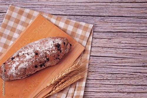 Fresh sourdough rests on wooden board, dusted with flour. Warm bread sits atop rustic cutting board. Wheat stalks lie beside it, woven mat below. Subtle shadow hints at soft, cozy kitchen light