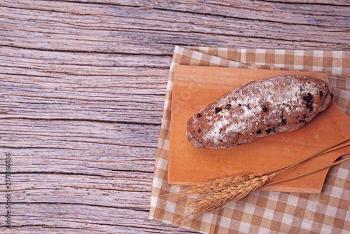Fresh sourdough rests on wooden board, dusted with flour. Warm bread sits atop rustic cutting board. Wheat stalks lie beside it, woven mat below. Subtle shadow hints at soft, cozy kitchen light