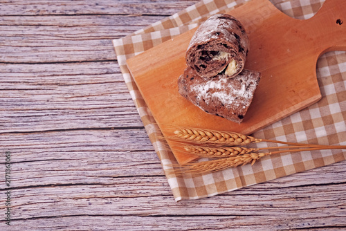 Sliced sourdough rests on a wooden board. Flour dusted loaf shows soft, chewy interior. Two wheat stalks add rustic charm. Checkered cloth beneath board enhances warmth. Ideal for food, bakery