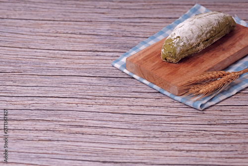Fresh sourdough dusted with flour sits on wooden board. Blue-checkered cloth beneath adds cozy contrast. Golden wheat stalks rest beside bread, hinting natural origins. Soft pastel wall behind creates