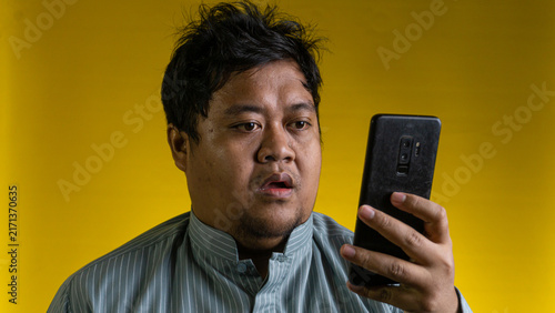Canvas Print A young Asian male displays a shocked facial expression while staring at a modern mobile device against a solid yellow background