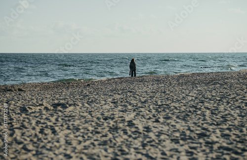 Woman standing and walking on empty sandy beach. Person alone at seashore with waves. Solitude, freedom and relaxation by the sea. Autumn vacation concept