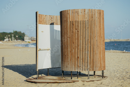 Wooden changing cabin on sandy beach. Open door of beach changing room hut with white interior. Sea and pier in background. Summer vacation and seaside leisure concept