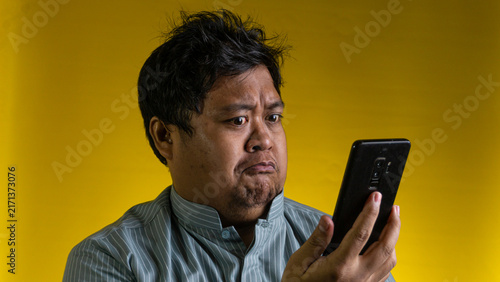 Bild auf Leinwand A young Asian male looks at a modern mobile device with a deep frown and a highly disappointed facial expression, standing against a solid yellow background