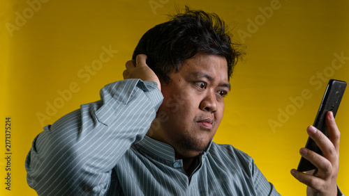 Foto A young Asian male scratches the back of the head while staring at a modern mobile device with a highly confused facial expression, set against a solid yellow background