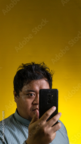 Foto A curious Asian male peeks cautiously from behind a vertically held smartphone against a solid yellow background, with the dark device obscuring exactly half of the face