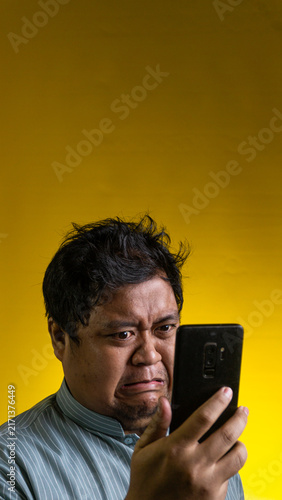 Fotografie A repulsed Asian male stares at a smartphone held in one hand against a bright yellow background, displaying a strong facial expression of intense disgust and rejection