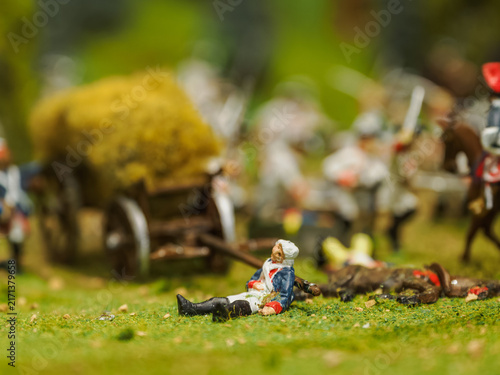 Fotografie A wounded soldier with a white wig and a blue coat in front of a wagon with straw on a battlefield