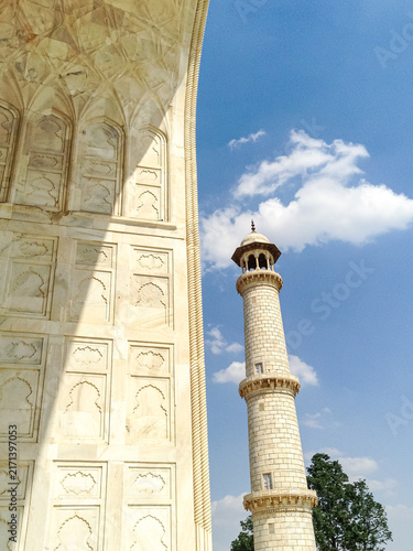 Close-up architectural view of a Taj Mahal minaret against blue sky, highlighting Mughal Islamic design, white marble detailing, and the monument's distinctive outward-leaning minaret structure.