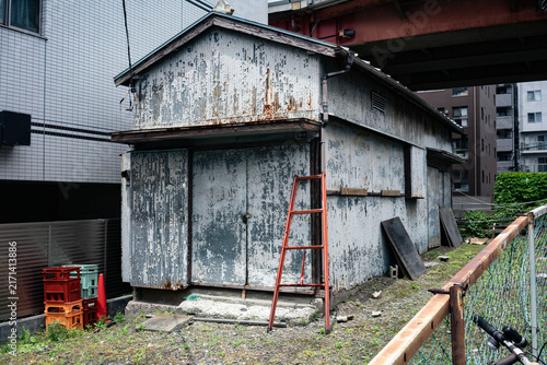 Fototapeta 塗装が剥げ落ちたトタン壁と赤い梯子が印象的な、都市の隙間に佇む古い小屋