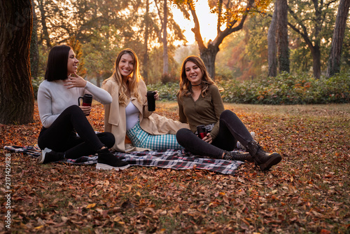 Three young women smile and enjoy warm drinks while relaxing on a picnic blanket in an autumn park filled with fallen leaves