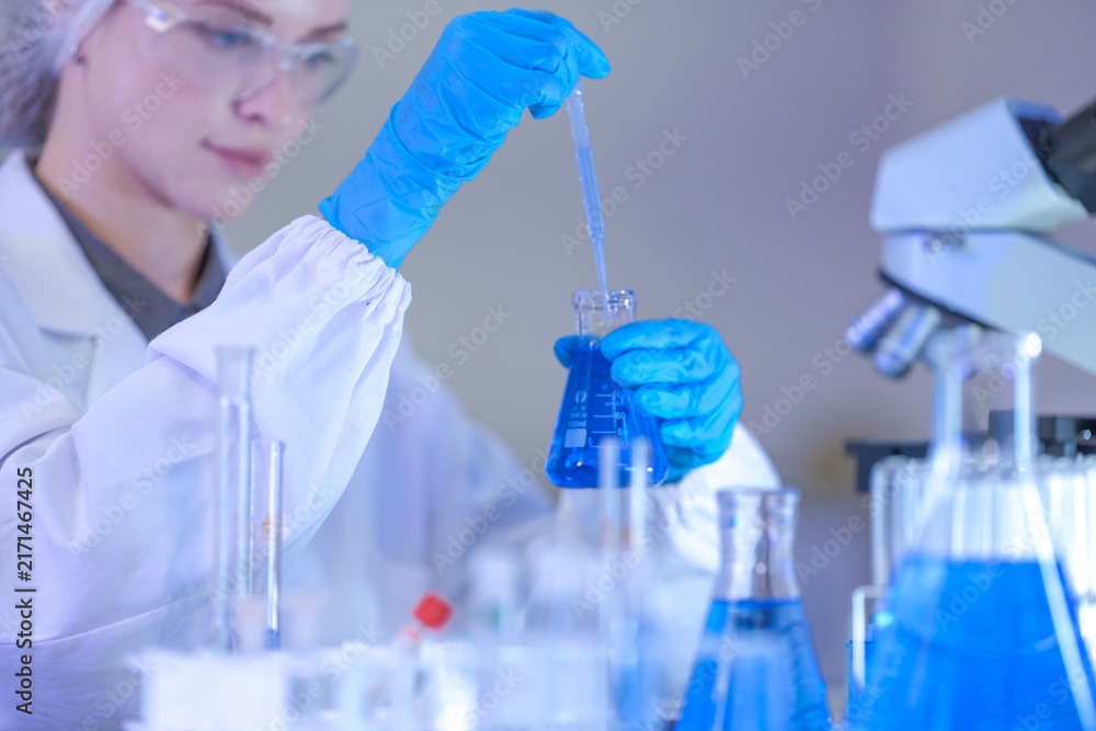 Fototapeta premium A woman researcher in white laboratory clothing, safety glasses and blue gloves holding the flask out beside the microscope at a brightly lit laboratory workbench.