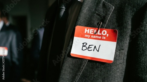 A bold red and white nametag prominently displayed on the lapel of a dark textured jacket, with a blurry figure wearing a similar tag in the background, suggesting a conference or networking