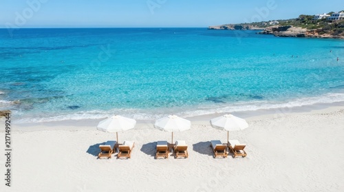 Aerial view of three sun loungers and white parasols on a pristine sandy beach by the clear turquoise waters of a summer coastline