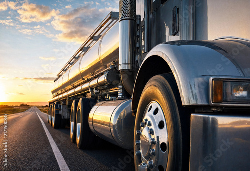 Polished stainless steel tanker truck driving along an open highway at sunset, with golden reflections, chrome details, crisp close-up focus, and blurred background.