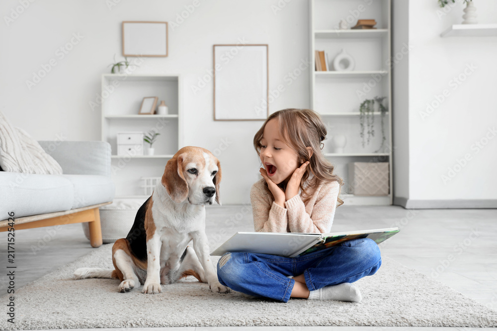 Fototapeta premium Surprised little girl with Beagle dog and book on floor in living room