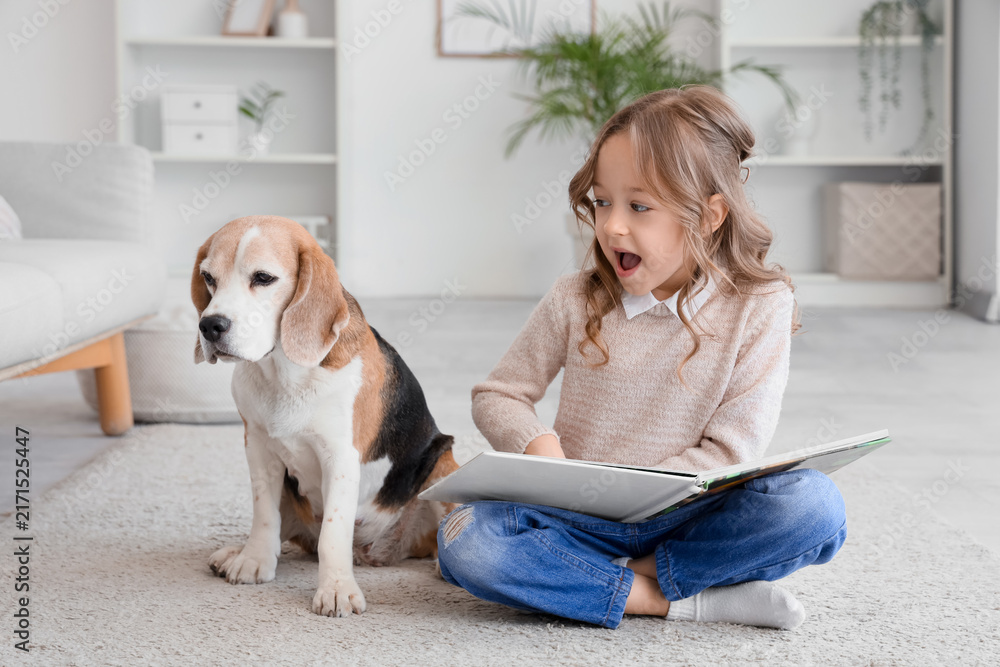 Fototapeta premium Cute little girl with Beagle dog reading book on floor in living room
