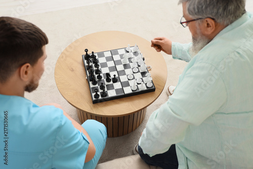 Young caregiver with senior man playing chess at home, back view