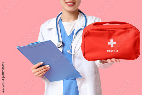 Asian female doctor with first aid kit and clipboard on pink background