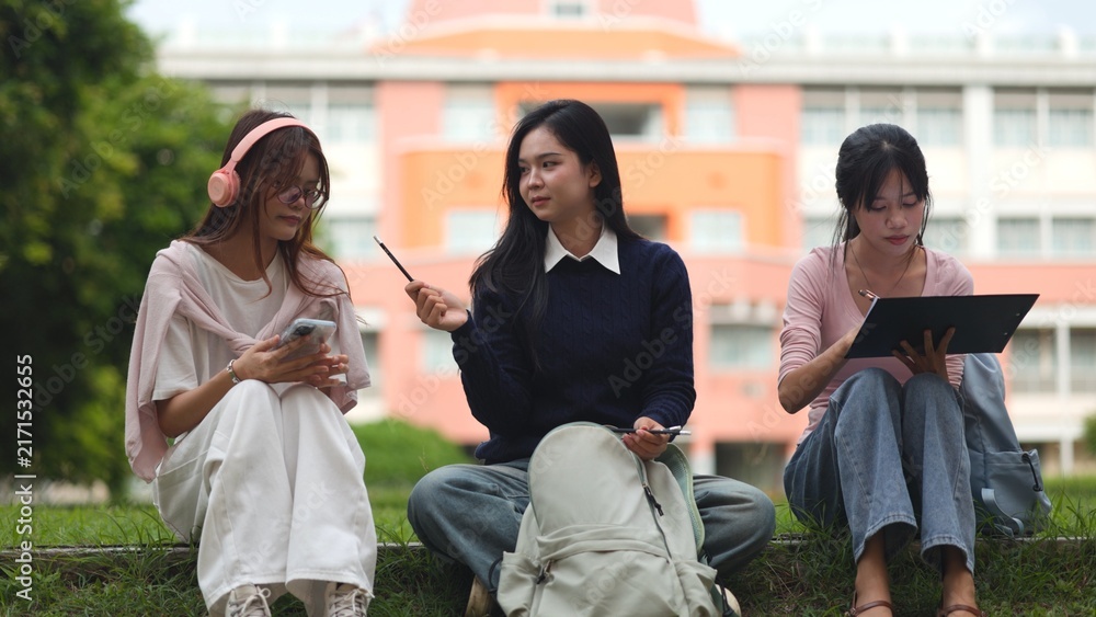 Obraz premium Asian university students relaxing and studying on green grass field with digital tablets and headphones. Modern higher education, Gen Z campus lifestyle, teamwork learning and youth concept.
