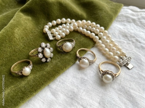 A collection of vintage pearl and diamond jewelry including rings and a multistrand pearl bracelet arranged on a green velvet fabric and white linen surface