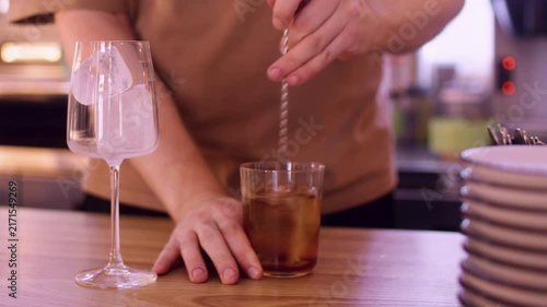 Refreshing drink making. Ice beverage. Bartender professionally stirring brown cold liquid with metal spoon in glass on pub counter.