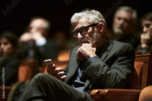 Mature man with grey hair and glasses looking at his smartphone while sitting in a dark theater