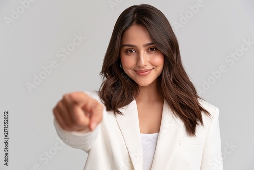 Smiling young woman in white blazer pointing forward with her finger against a grey background