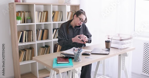 Corporate routine. Office makeup. Female manager talking on smartphone applying face powder at desk with laptop for business paperwork indoor.