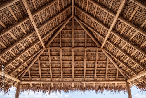 Low angle view of a thatched roof structure with wooden beams against a blue sky